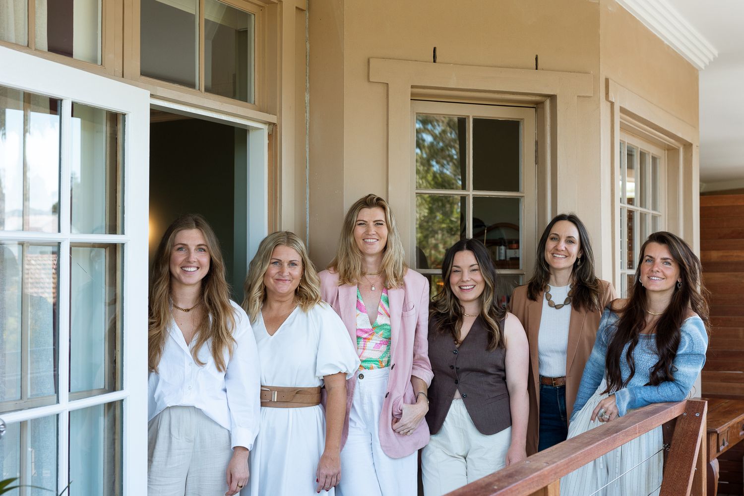 Seven women smiling together on a sunlit porch, dressed in elegant casual outfits for a social gathering.