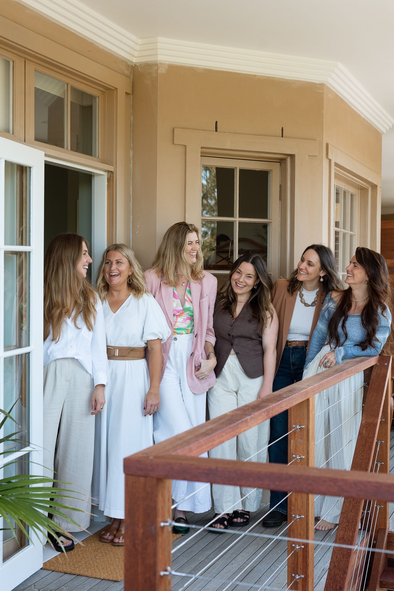 Six women laughing and posing together on a wooden verandah of a classic home on a sunny day.