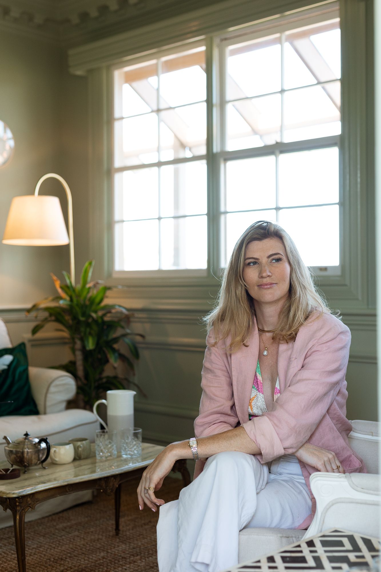 Woman in pink blazer and white pants sits in elegant room with large windows and soft lighting.
