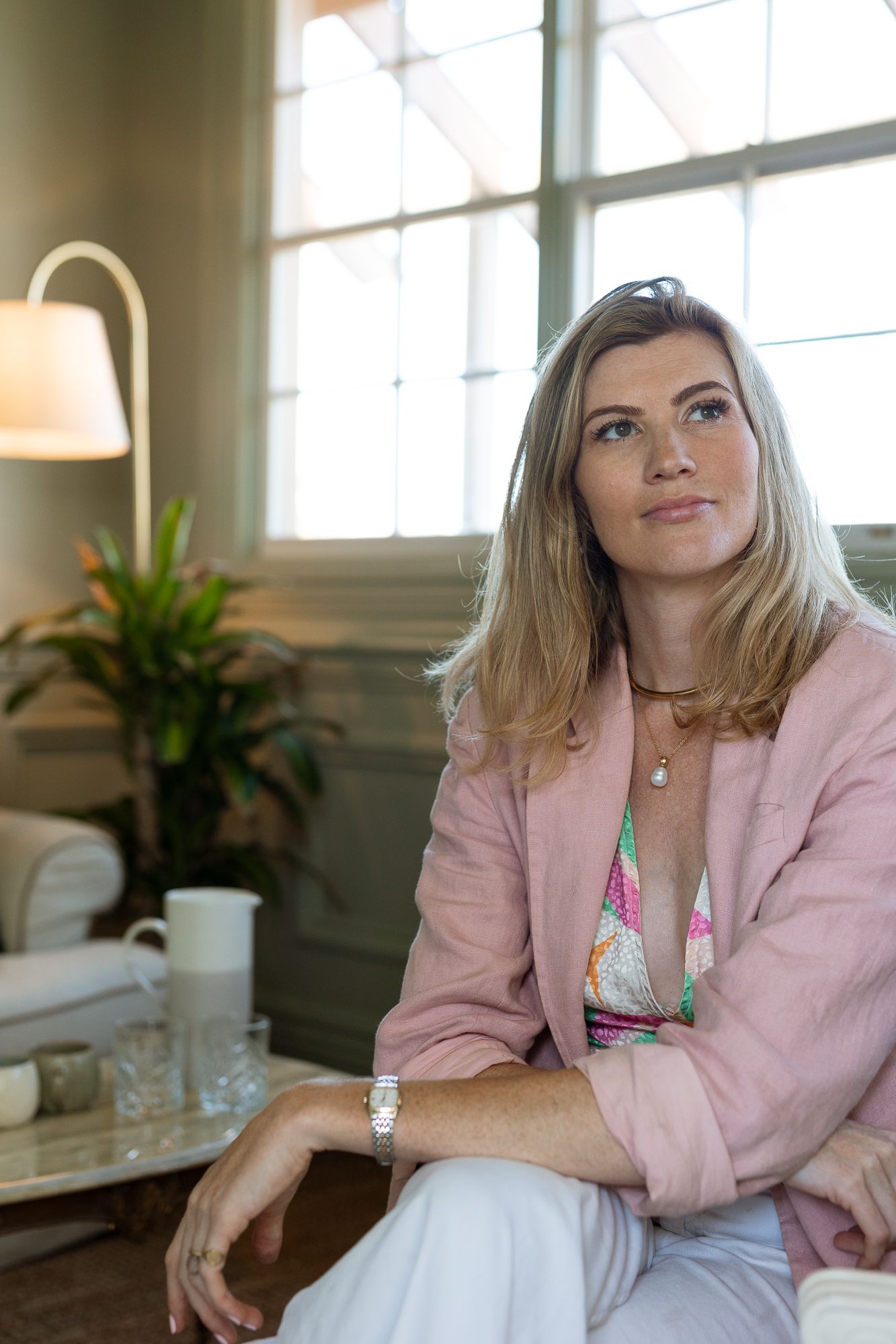 Blonde woman in pink blazer and colorful top sits thoughtfully in elegant living room with natural light.