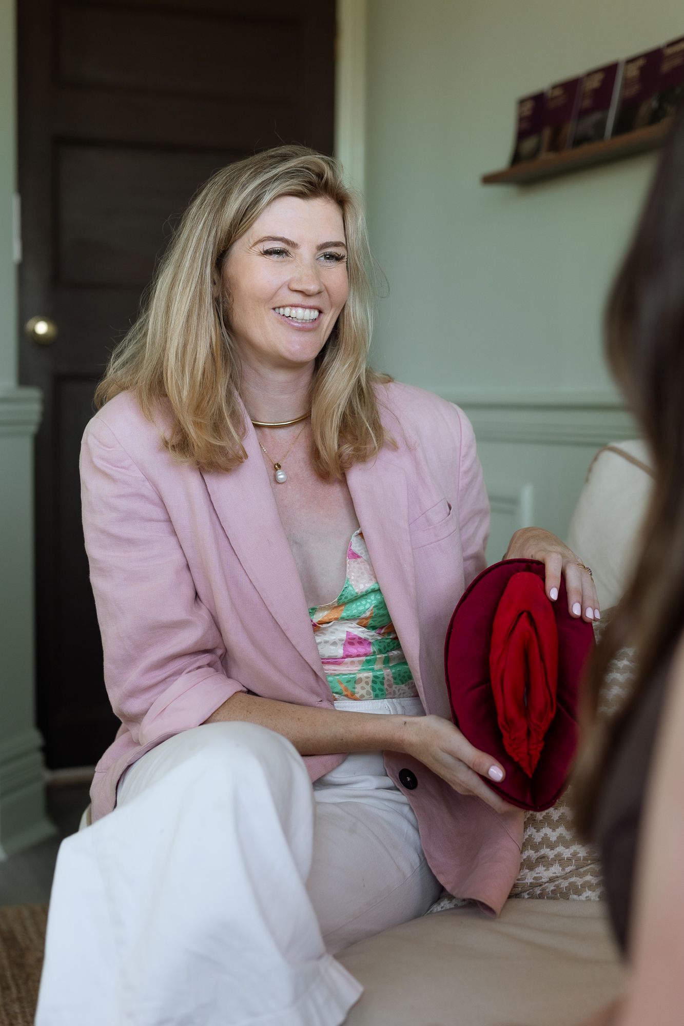 Smiling blonde woman in pink blazer holding red hat, seated indoors in bright casual setting.