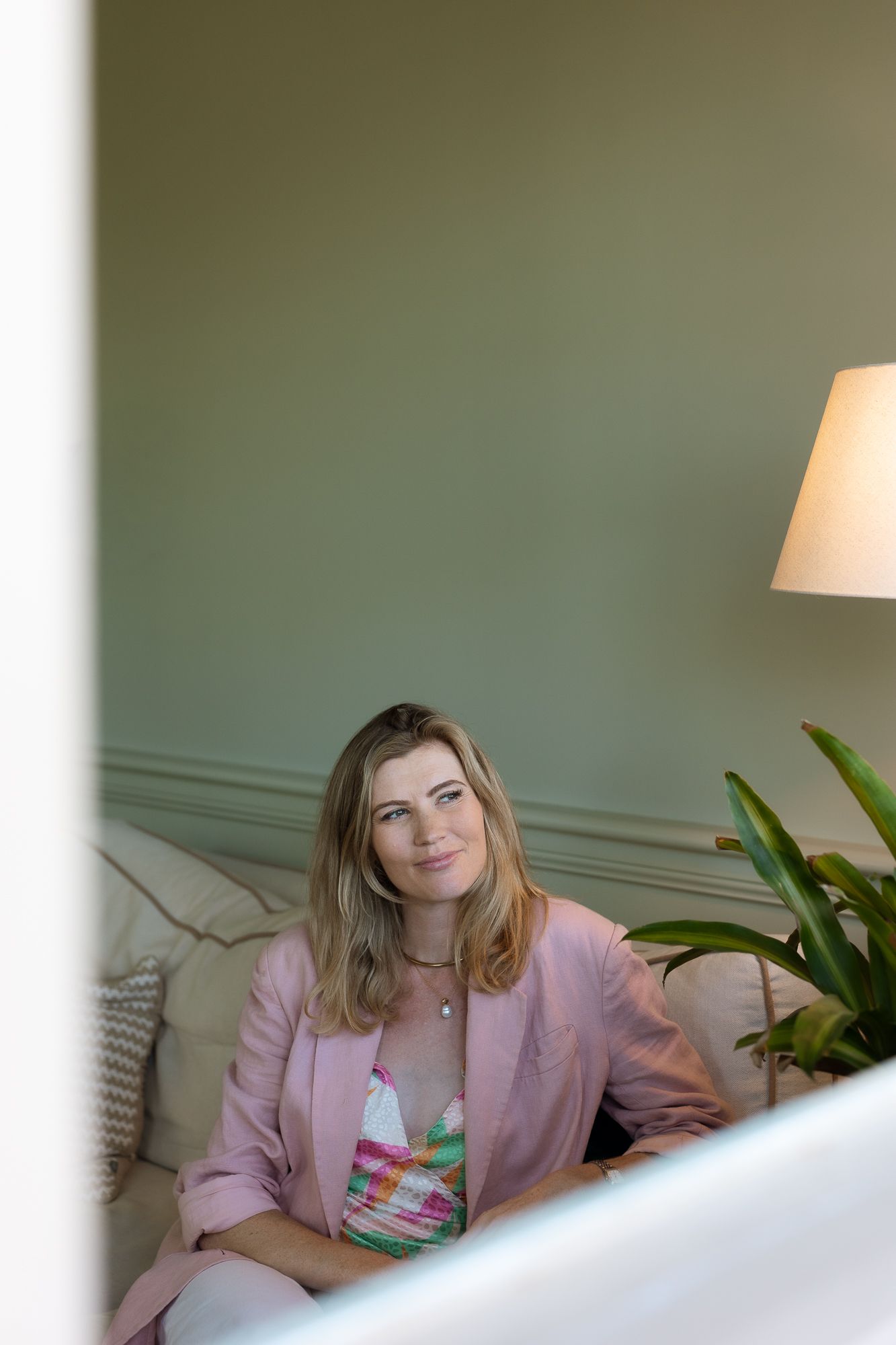 Woman in pink blazer sitting on a beige sofa against a sage green wall with a lamp and plant nearby.
