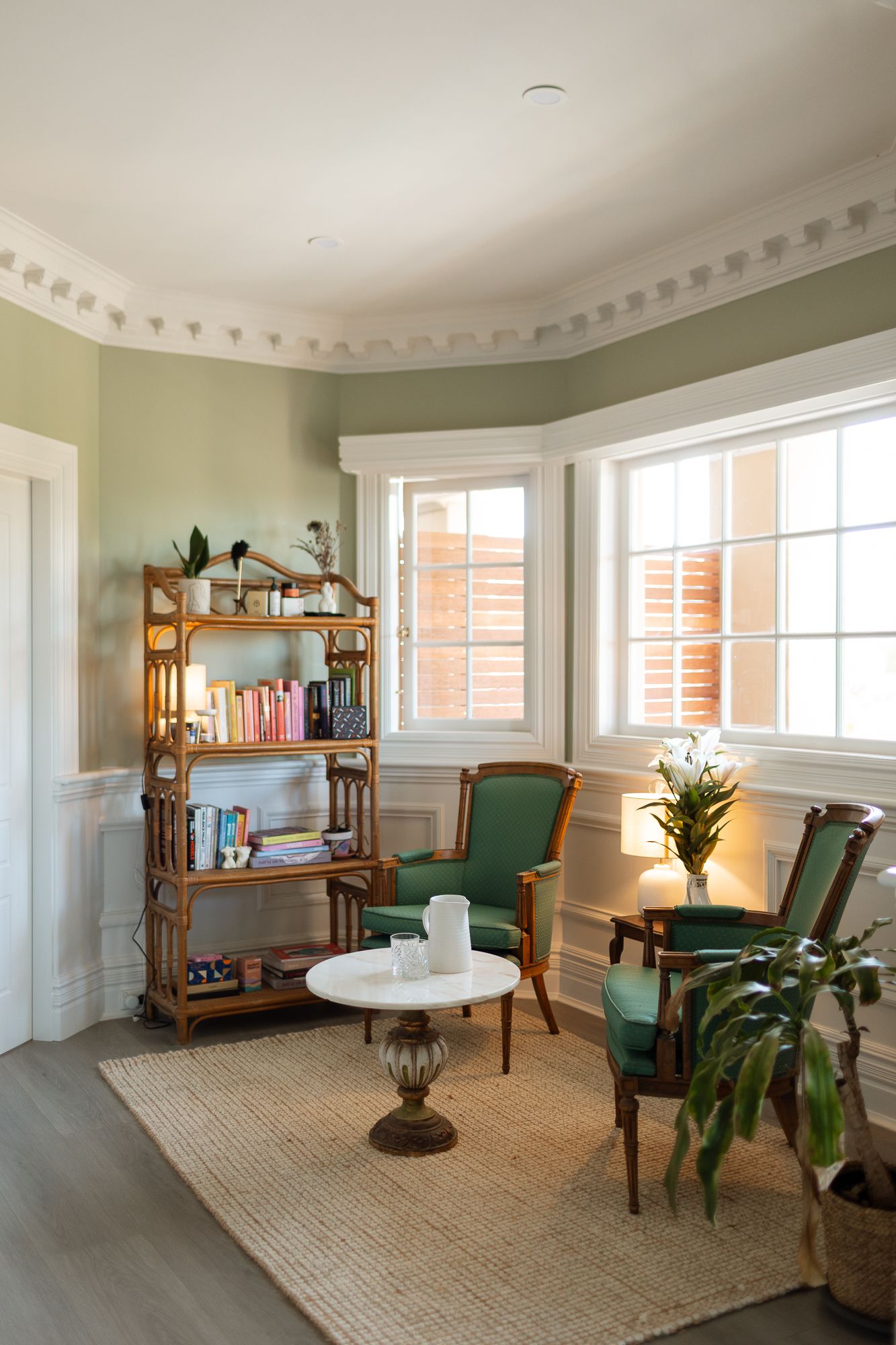 Cozy reading nook with green armchairs, wooden bookshelf, small white table, and bay windows in a sage green room.