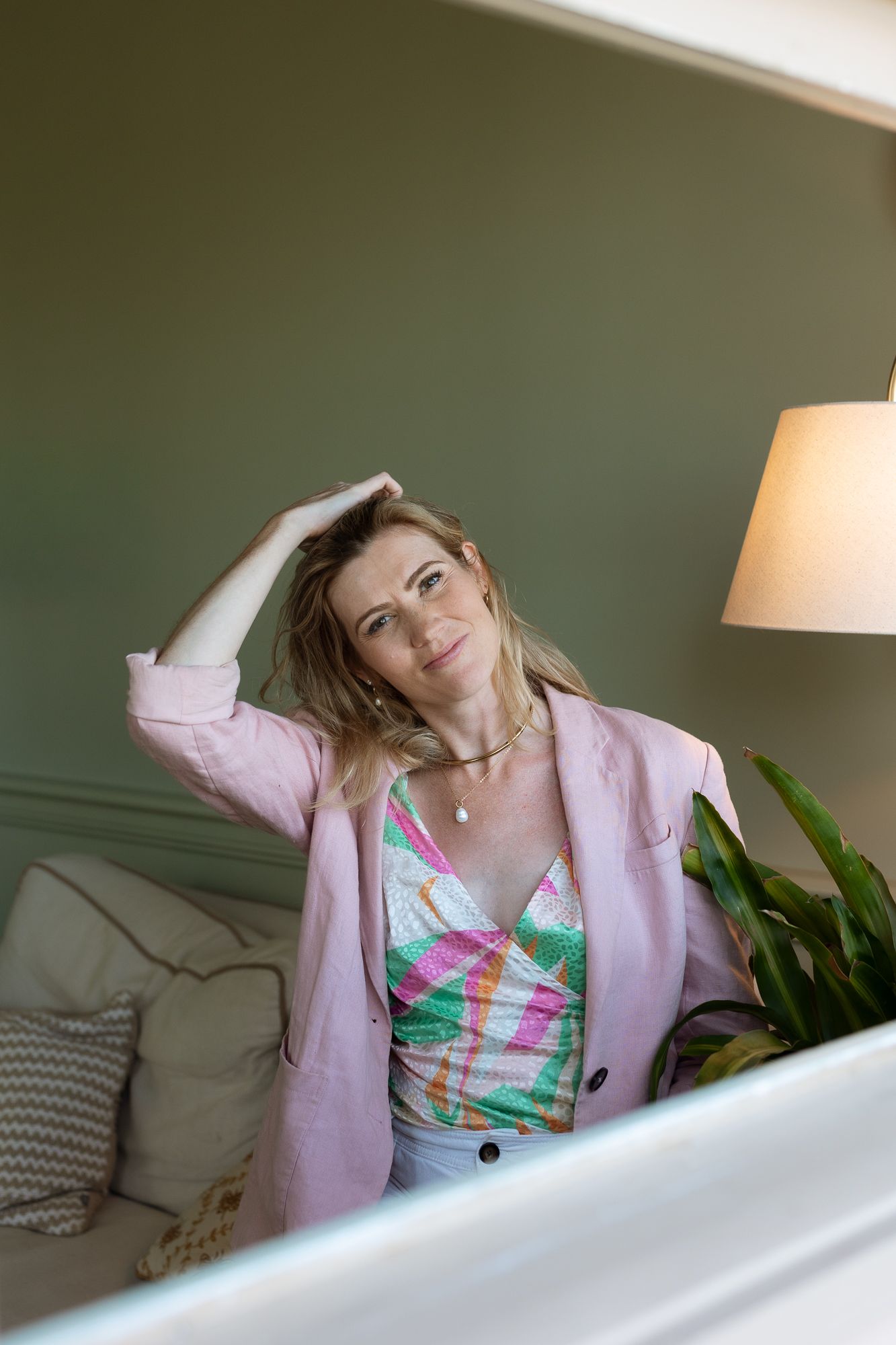 Woman in pink blazer and floral top poses thoughtfully beside a lamp and green plant in a cozy room.