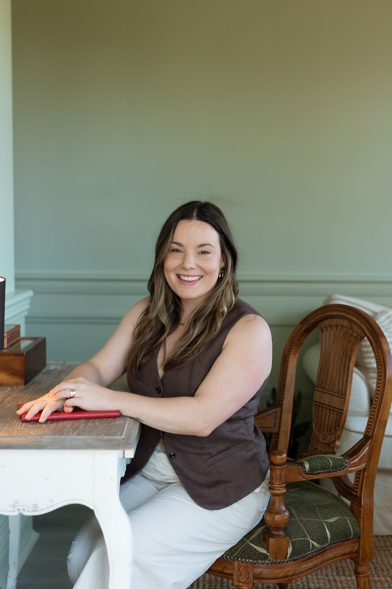 Smiling woman in brown vest sits at rustic white table beside wicker chair in sage green room.