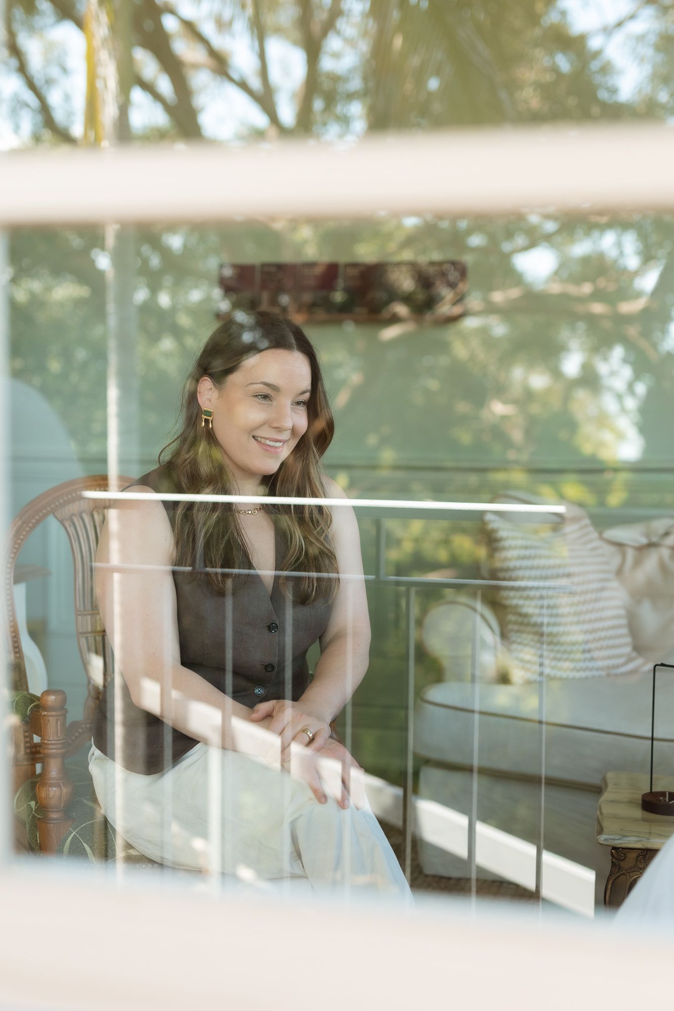 Smiling woman with long hair sitting on an outdoor balcony chair, surrounded by lush green trees.