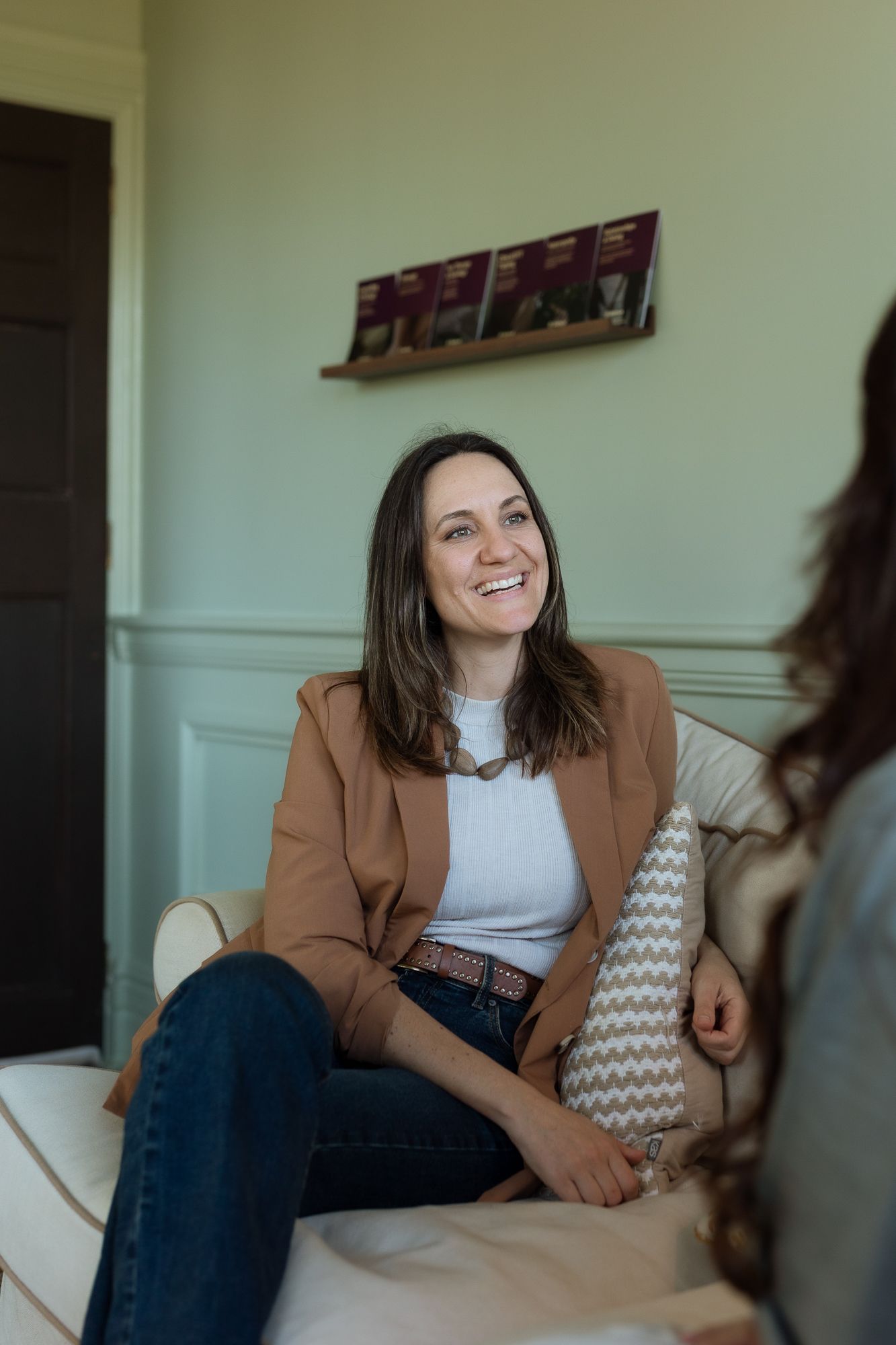 Smiling woman in brown blazer sitting on white sofa holding decorative pillow in bright modern room.