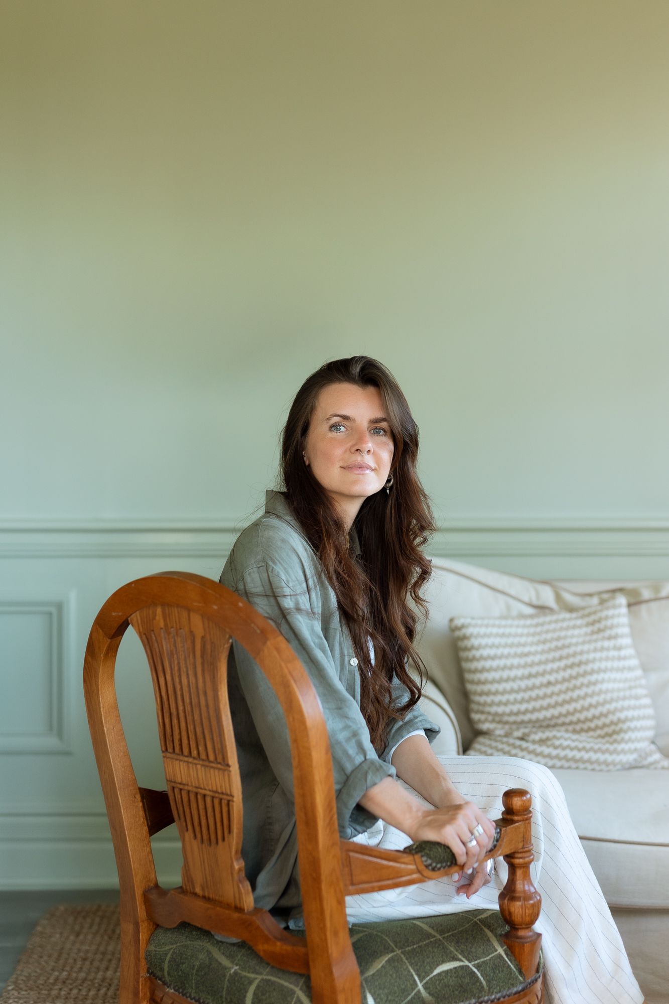 Woman with long dark hair sits on wooden chair in sage green room wearing linen shirt and white pants.