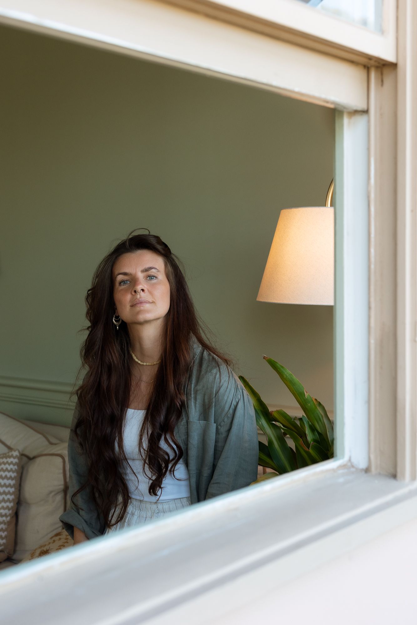 Woman with long dark hair sitting by a window in a cozy green room with a lamp and plant.