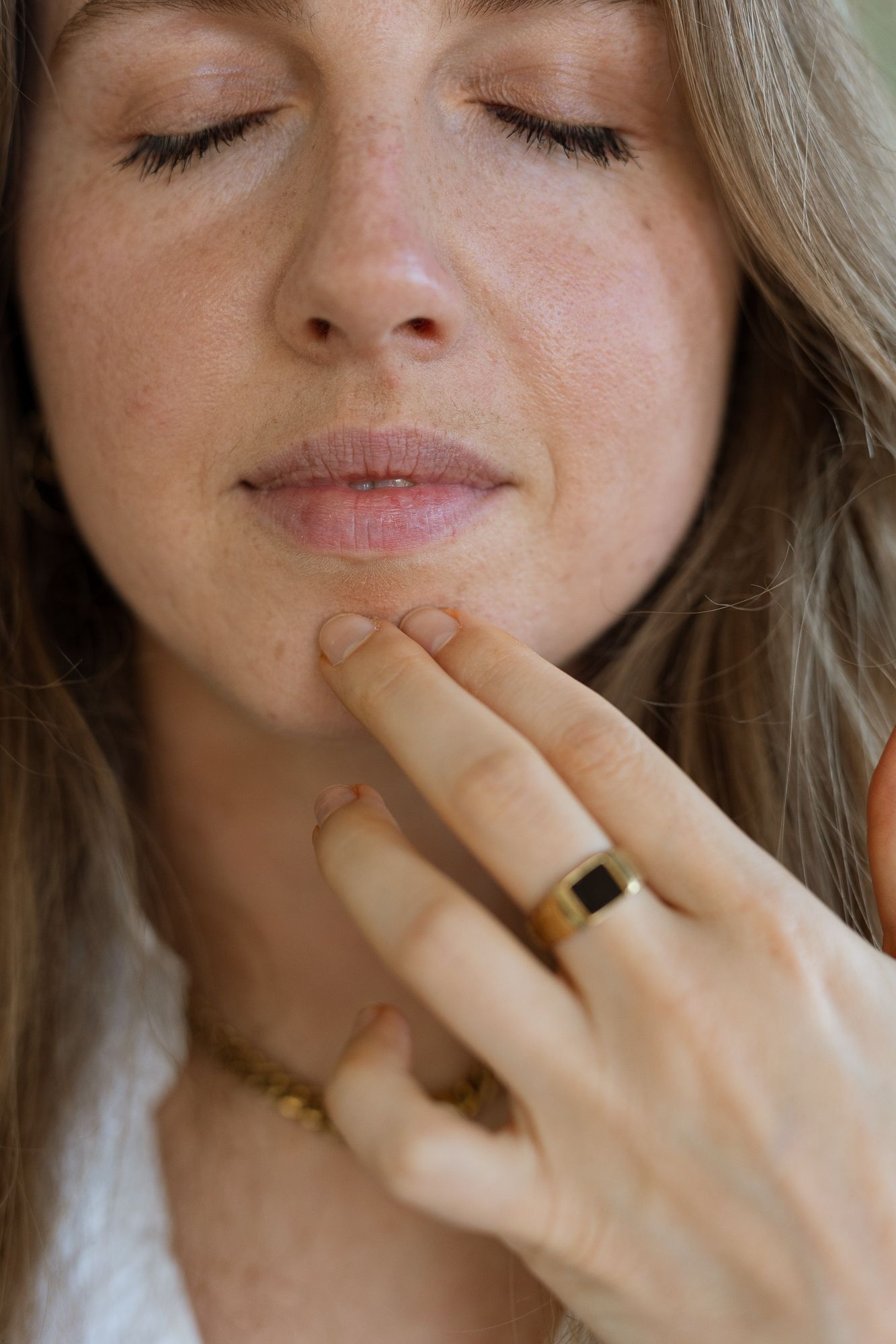 Close-up of a woman with closed eyes touching her chin, wearing a gold ring and delicate necklace.