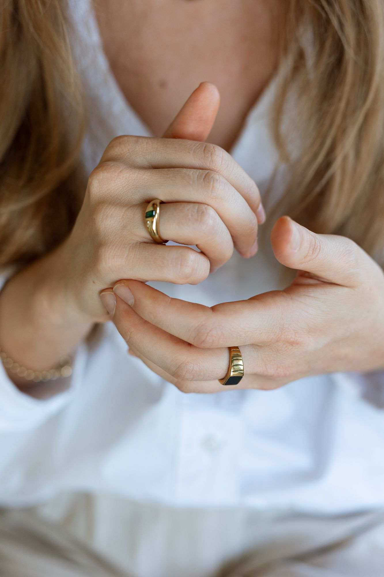 Close-up of hands wearing gold rings, one with a green gemstone, clasped together over a white shirt.
