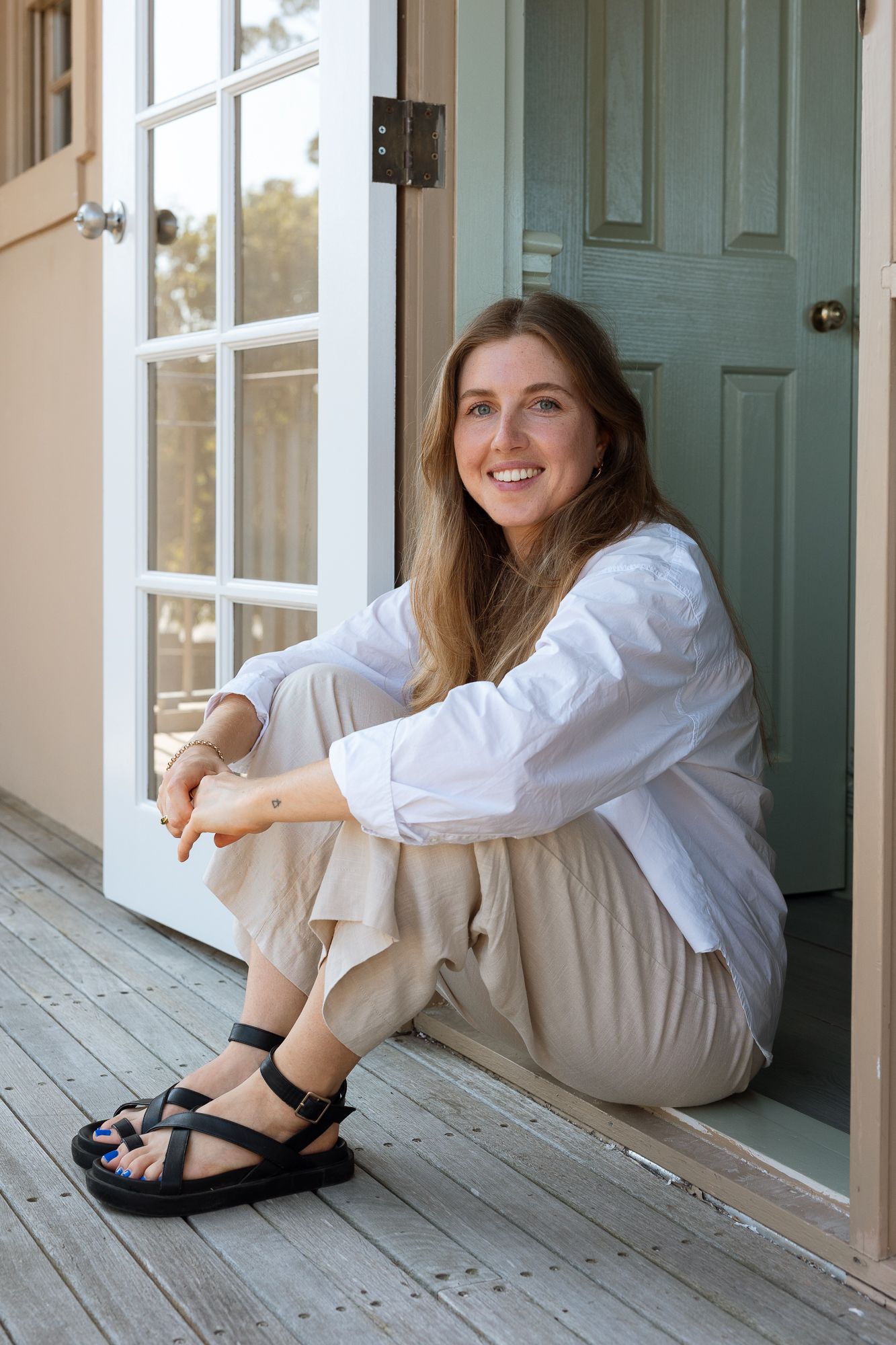 Smiling woman in white shirt and beige pants sitting on wooden porch steps by a sage green door wearing black sandals.