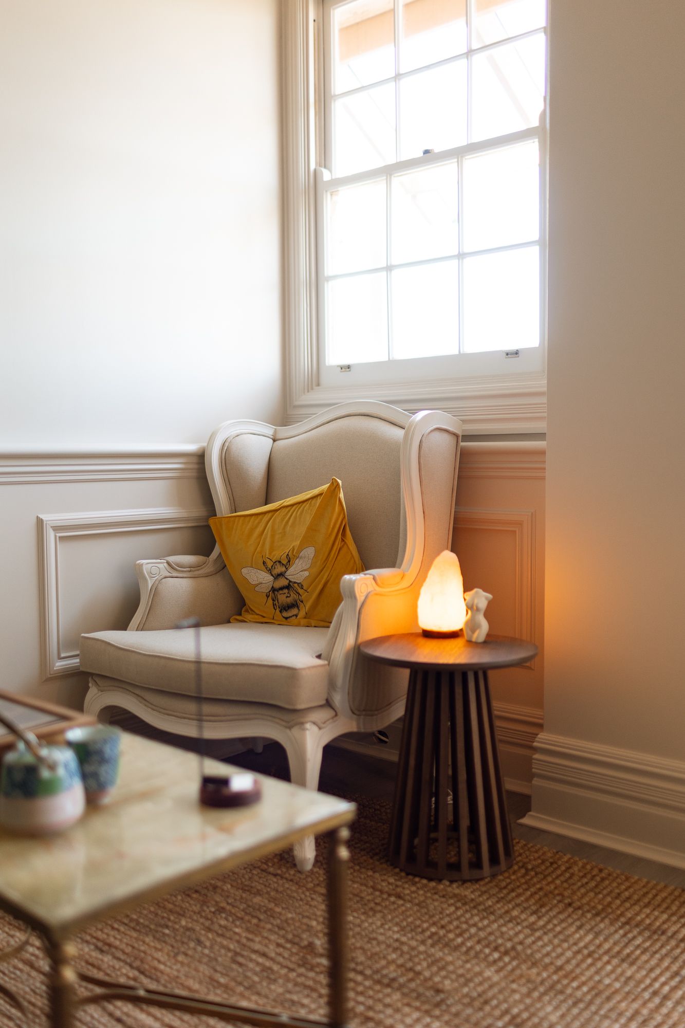 Cozy reading nook with a vintage wingback chair, bee pillow, glowing salt lamp on side table, and natural light from window.