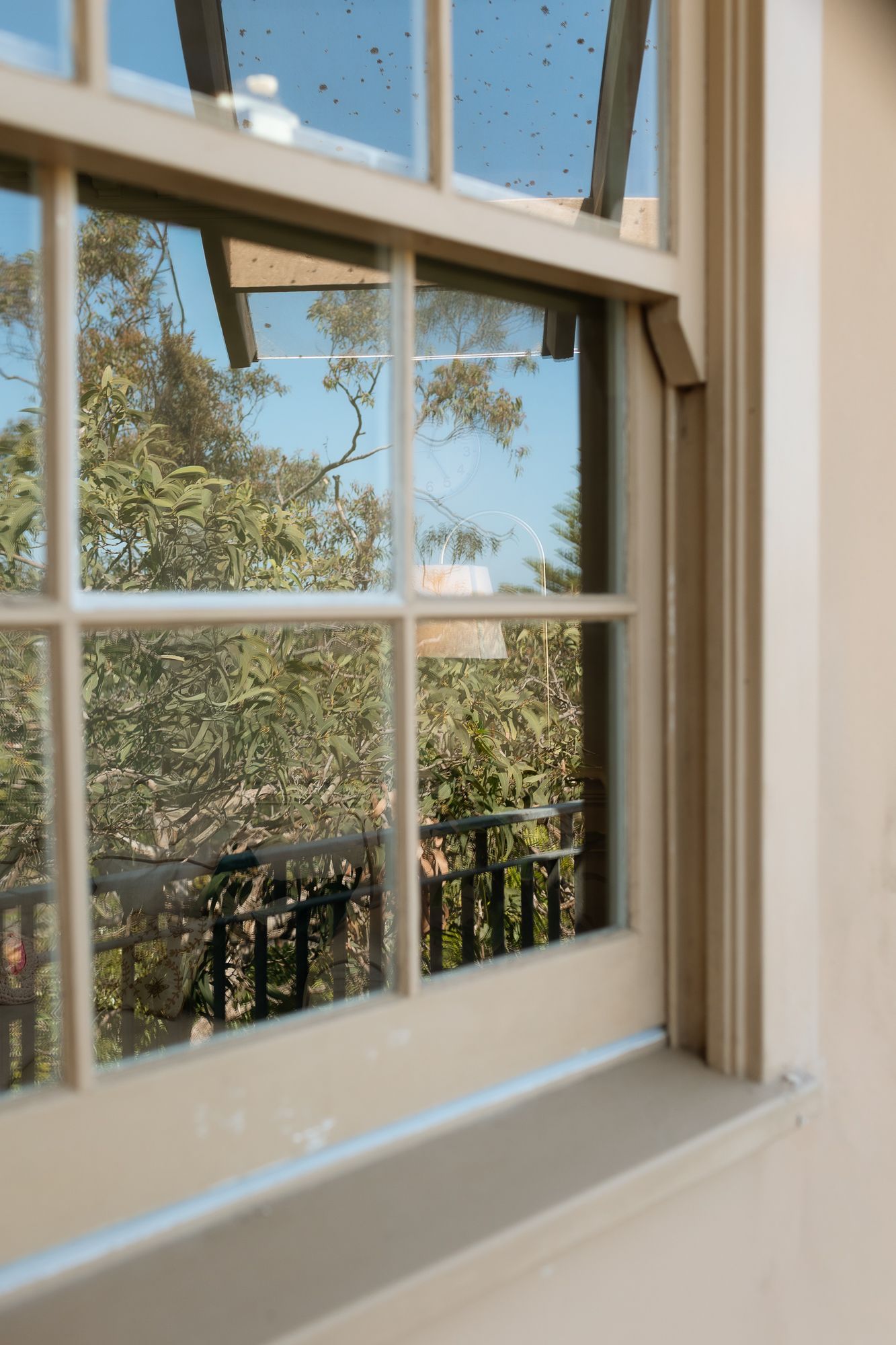 White timber double-hung window with grid panes overlooking a balcony with iron railing and lush green trees beyond.