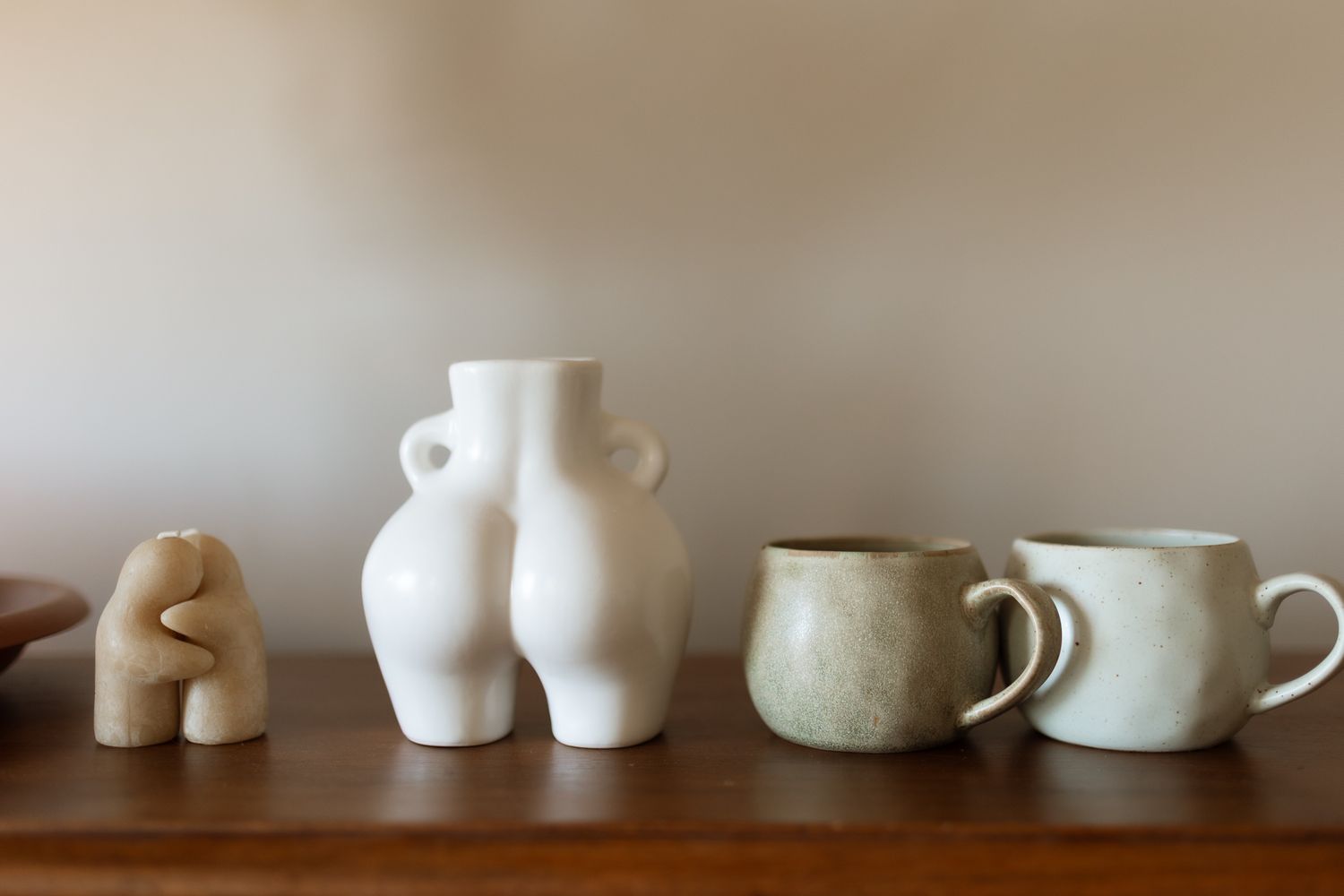 Minimalist shelf display with a sculptural wood figurine, white ceramic vase, and two speckled stoneware mugs.