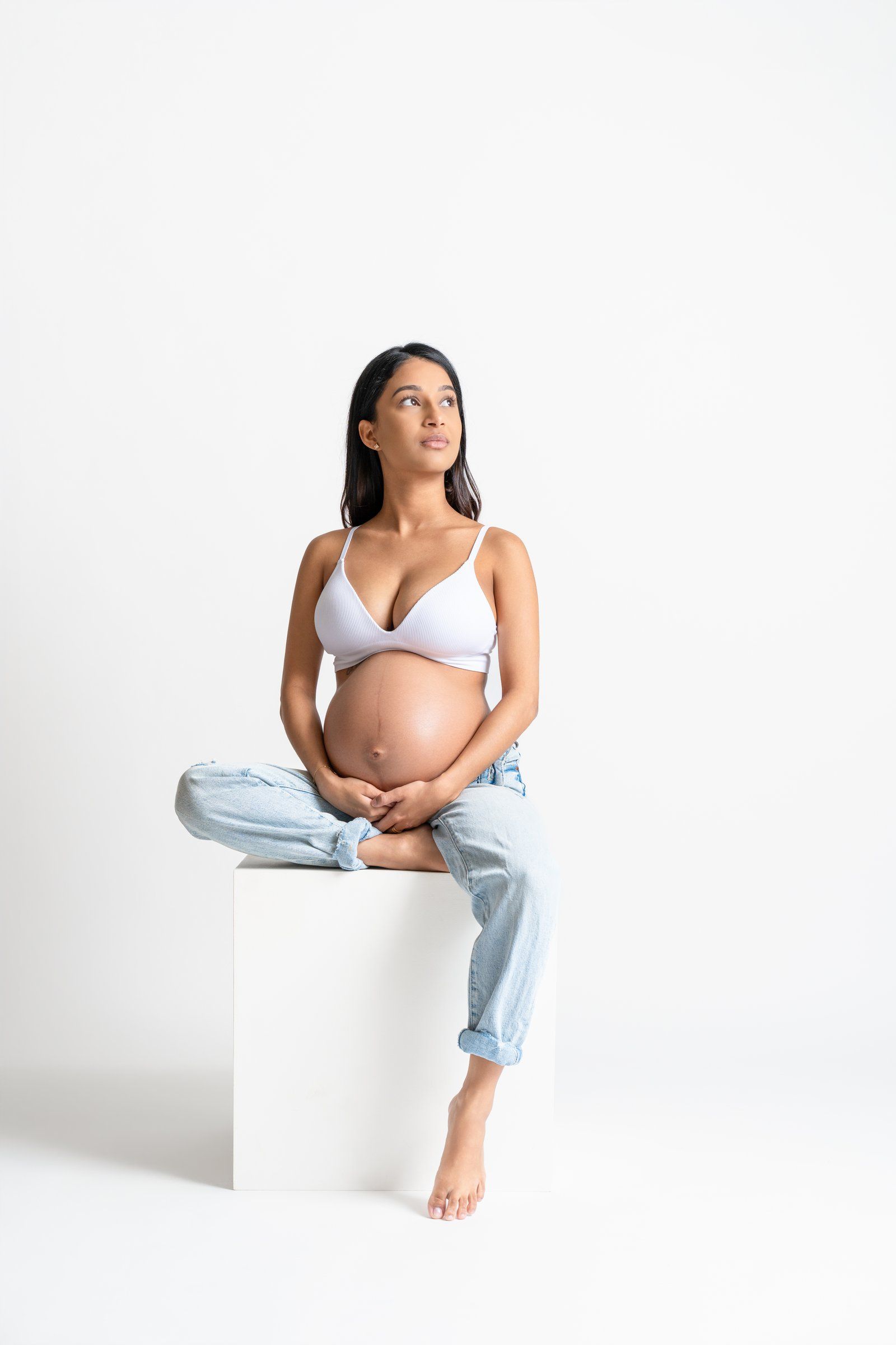 Pregnant woman in white bra and jeans sitting on white cube, cradling baby bump, looking up thoughtfully.