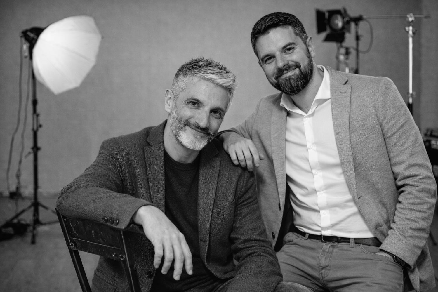 Two smiling bearded men posing together in black and white studio photo with lighting equipment.