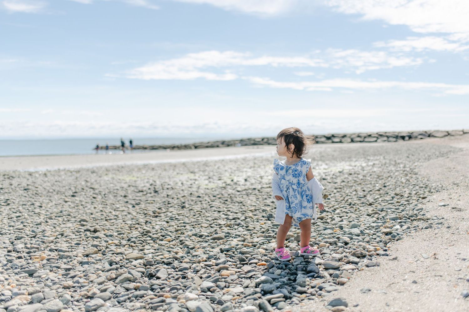 Young girl in floral dress and pink boots standing on a rocky pebble beach under a cloudy sky.
