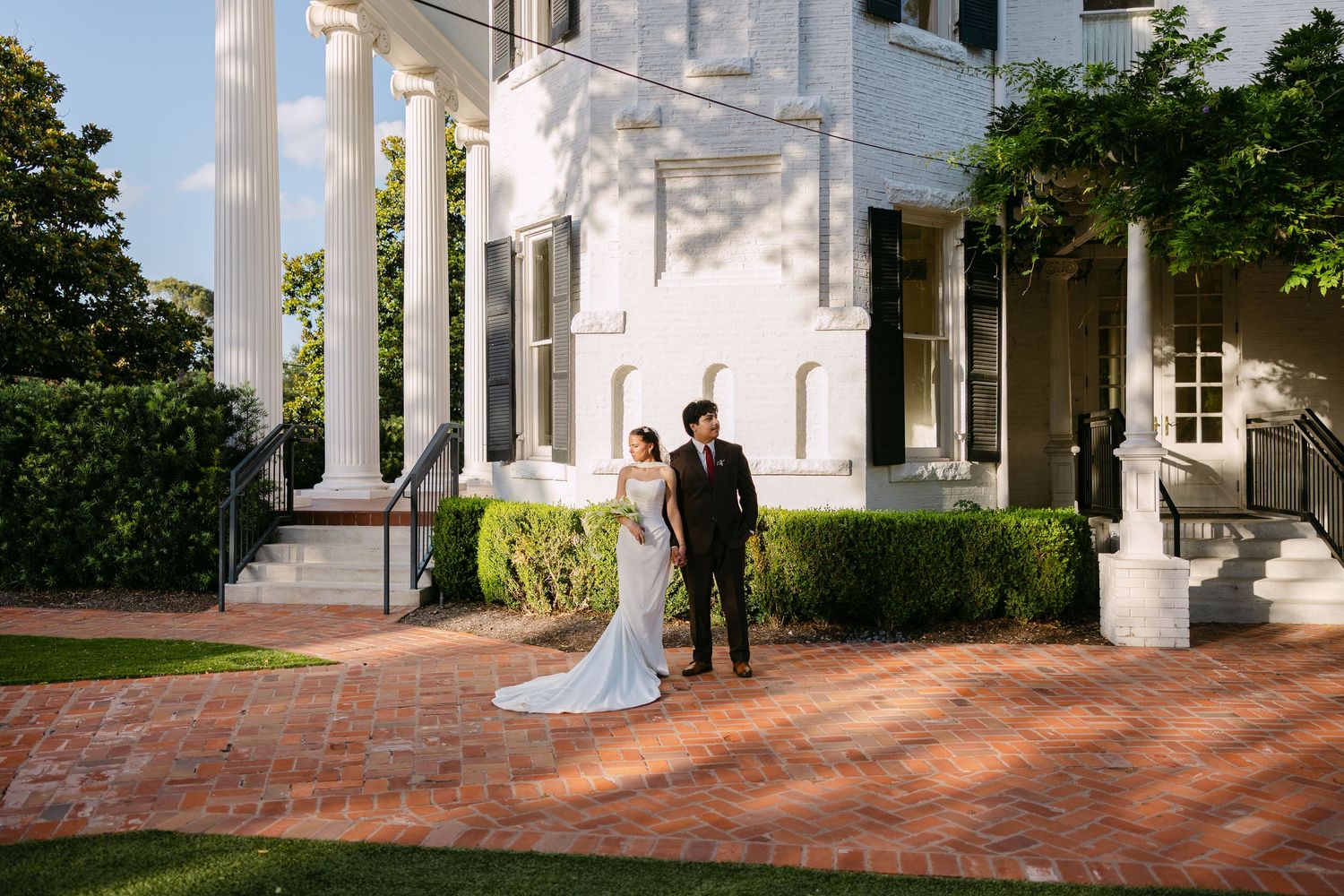 Bride and groom pose on brick pathway outside elegant white colonial mansion with tall columns on wedding day.