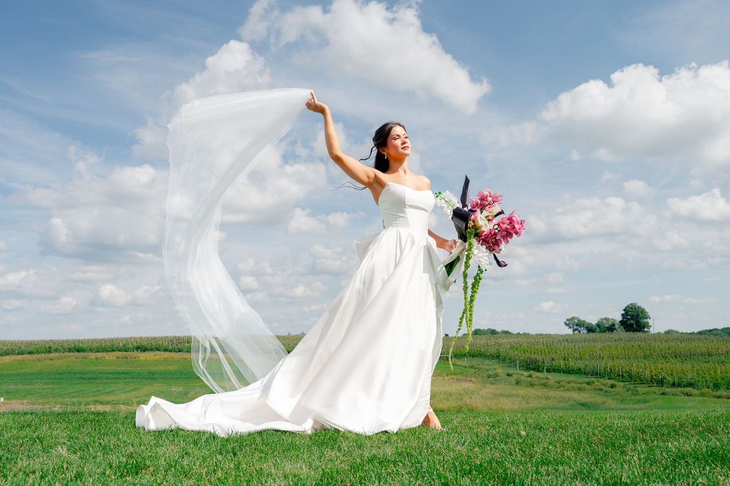 Bride in white gown holding veil and bouquet outdoors on green field with blue sky.