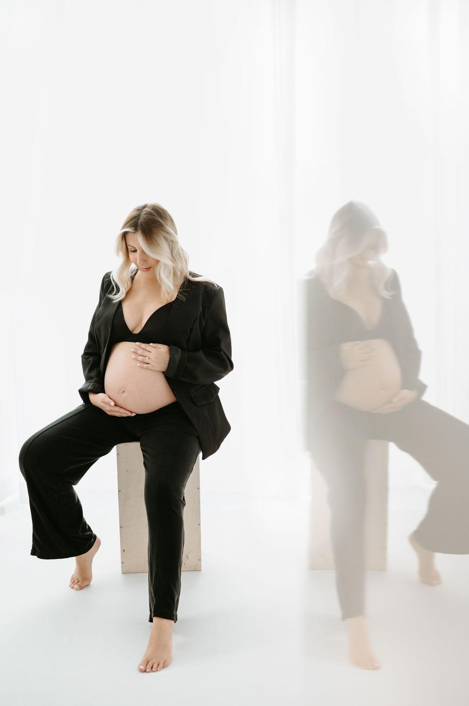 Pregnant woman in black blazer and pants sits barefoot, cradling her baby bump, with a soft reflection beside her.