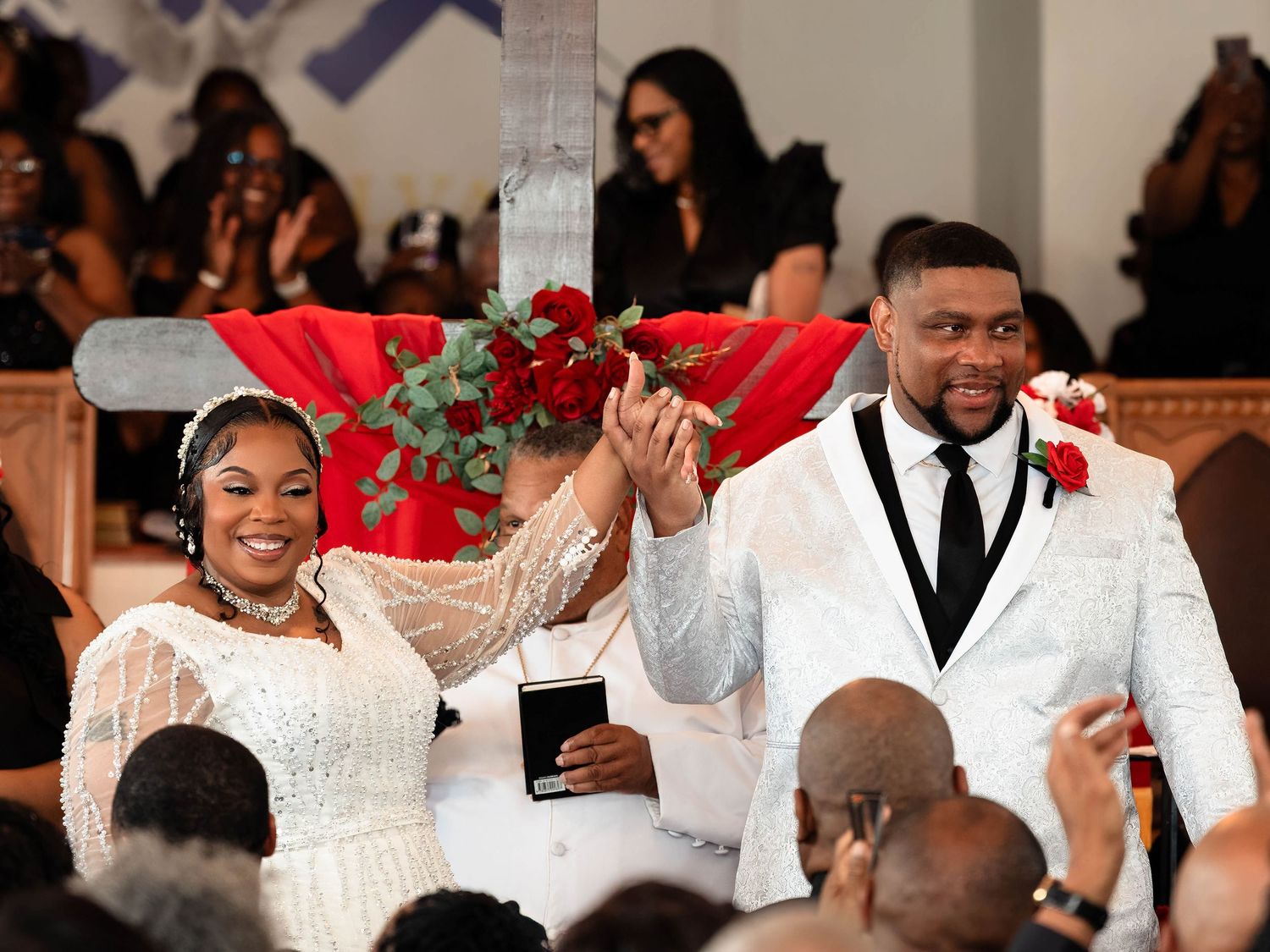 Joyful newlyweds celebrate at church wedding, bride in white gown holding red roses, groom in white suit raising hands.