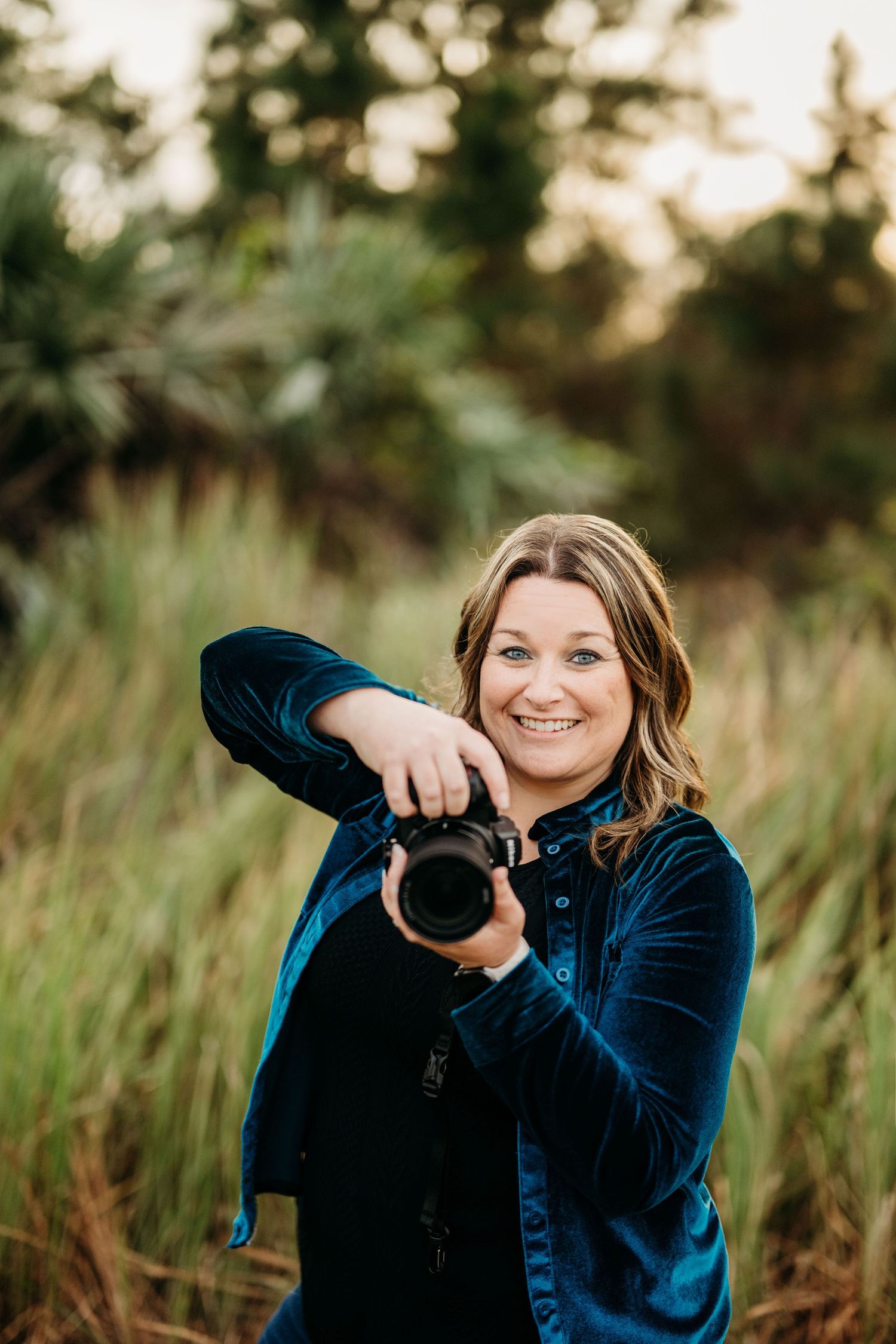 A photographer wearing a blue velvet jacket holds a camera while standing in tall grass at sunset.
