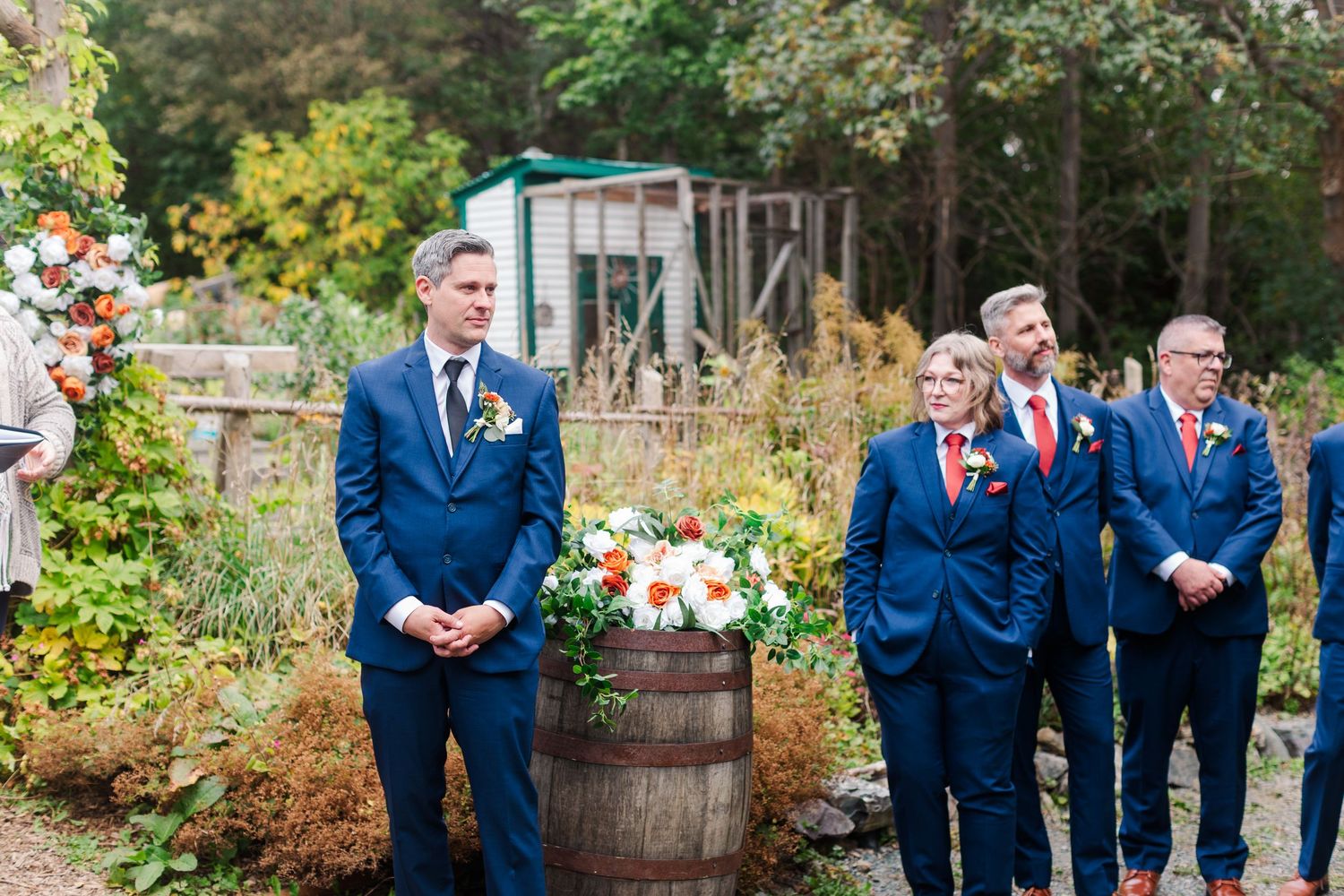 Groomsmen in navy suits stand in a row at an outdoor garden wedding ceremony beside a wooden barrel topped with a floral arrangement.