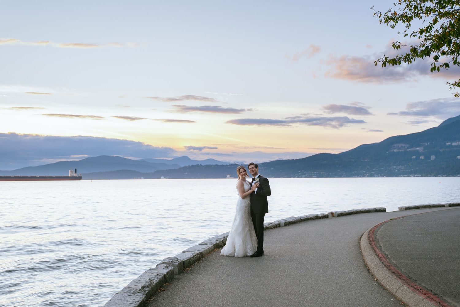Bride and groom embrace on a waterfront path at sunset with mountains and calm water in the background.