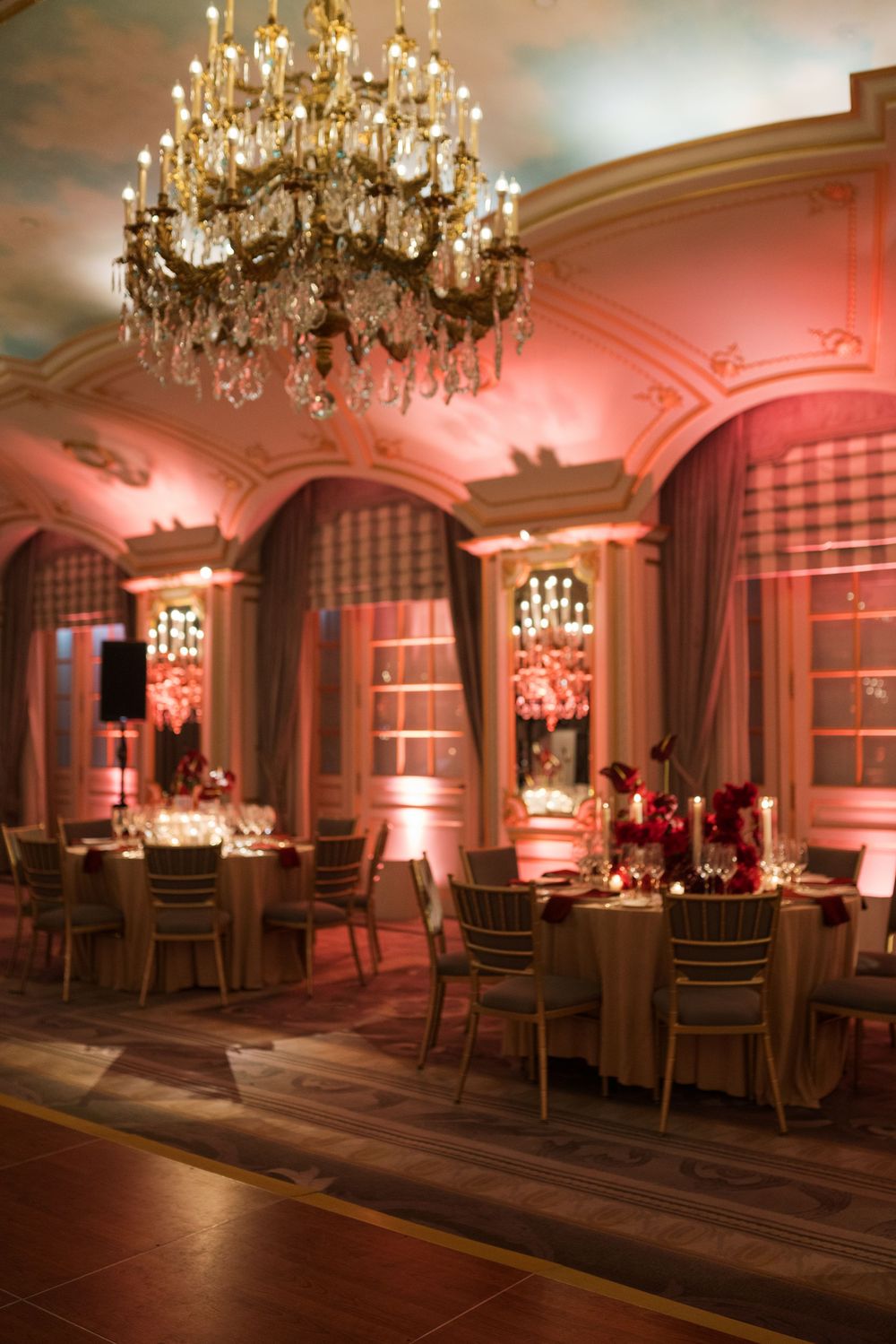 Elegant ballroom with crystal chandelier, pink uplighting, round dining tables, and ornate arched windows for a luxurious event.