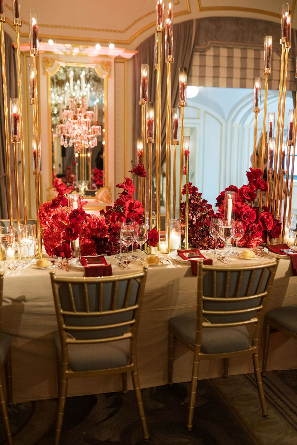 Elegant dining table with red floral centerpieces, gold candelabras, and mirrored walls in a luxurious romantic setting.