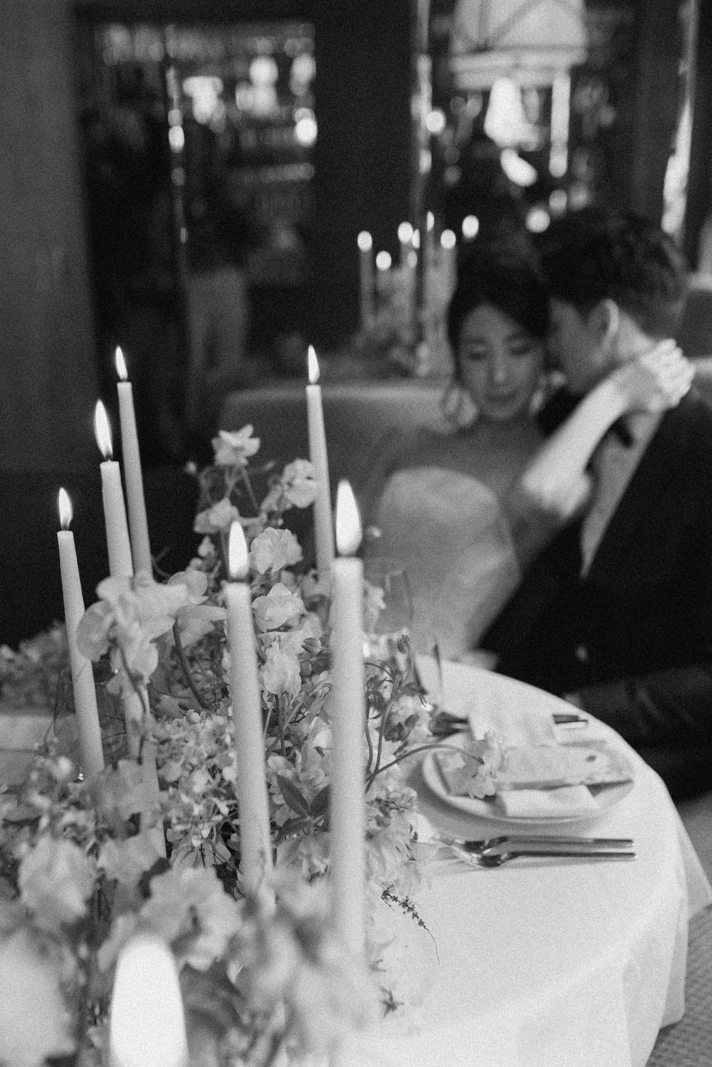 Black and white wedding photo of couple embracing behind elegant candlelit table with floral centerpiece.