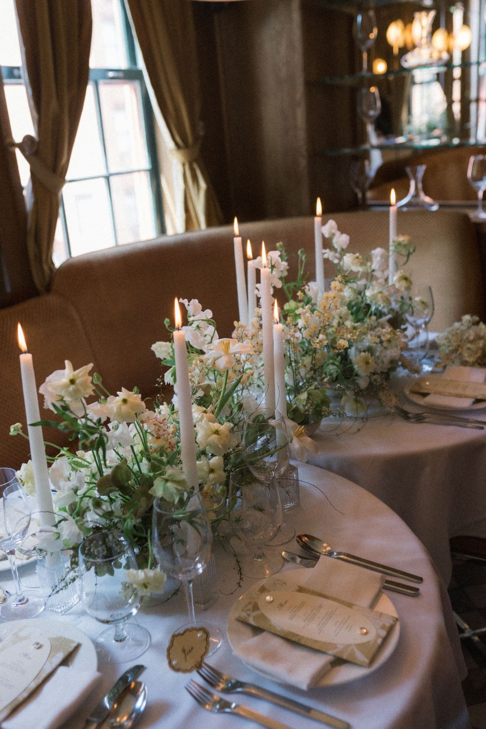 Elegant wedding table setting with white taper candles, soft floral arrangements, and fine silverware on a draped table.