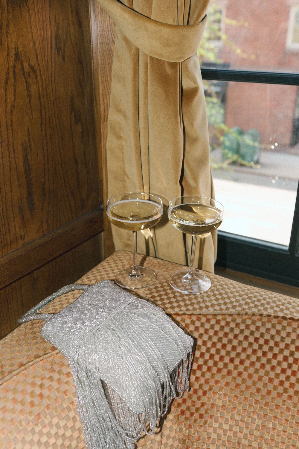 Two elegant champagne glasses with white wine beside a silver clutch purse on a checkered ottoman near a curtained window.