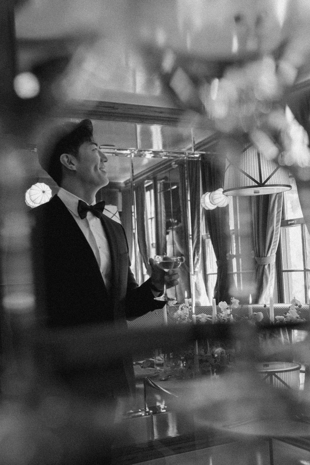 Black and white photo of a smiling man in a tuxedo holding a cocktail glass at an elegant formal event.