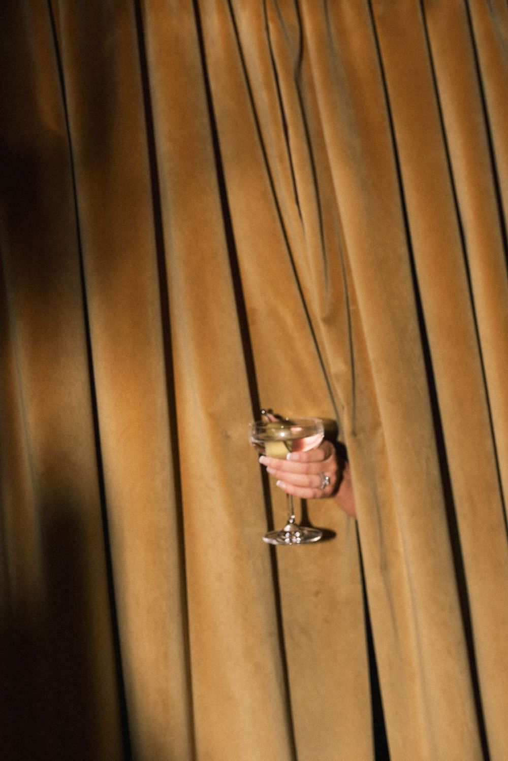A hand holding a martini glass emerges through golden velvet curtains in a dramatic theatrical setting.