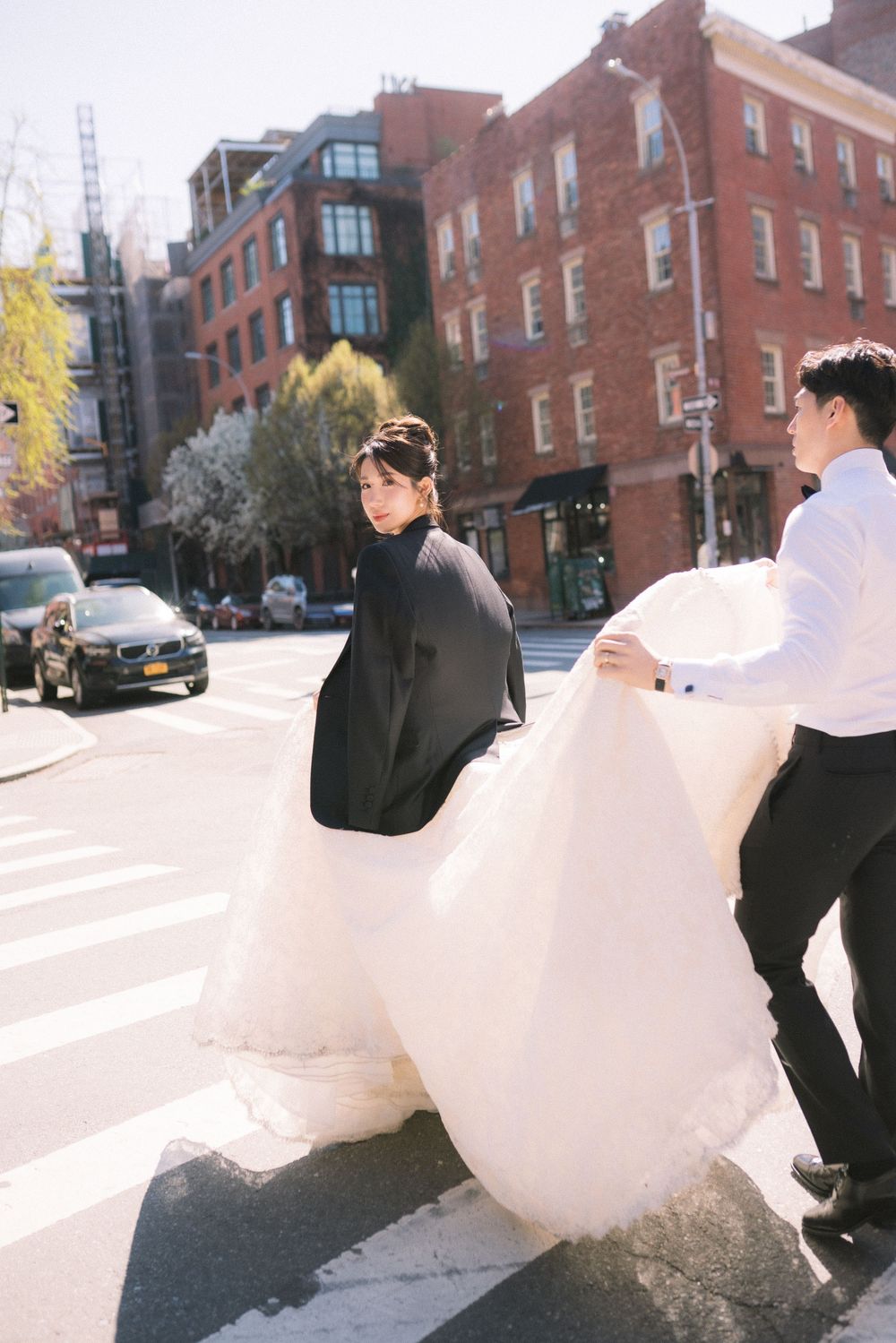 Bride in black jacket and white gown crosses NYC street as groom holds her train, with brick buildings in background.