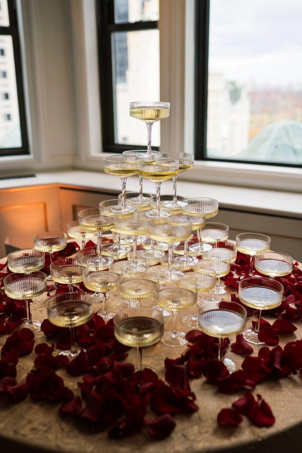 Elegant champagne tower with crystal coupe glasses filled with champagne, surrounded by red rose petals on a table near windows.