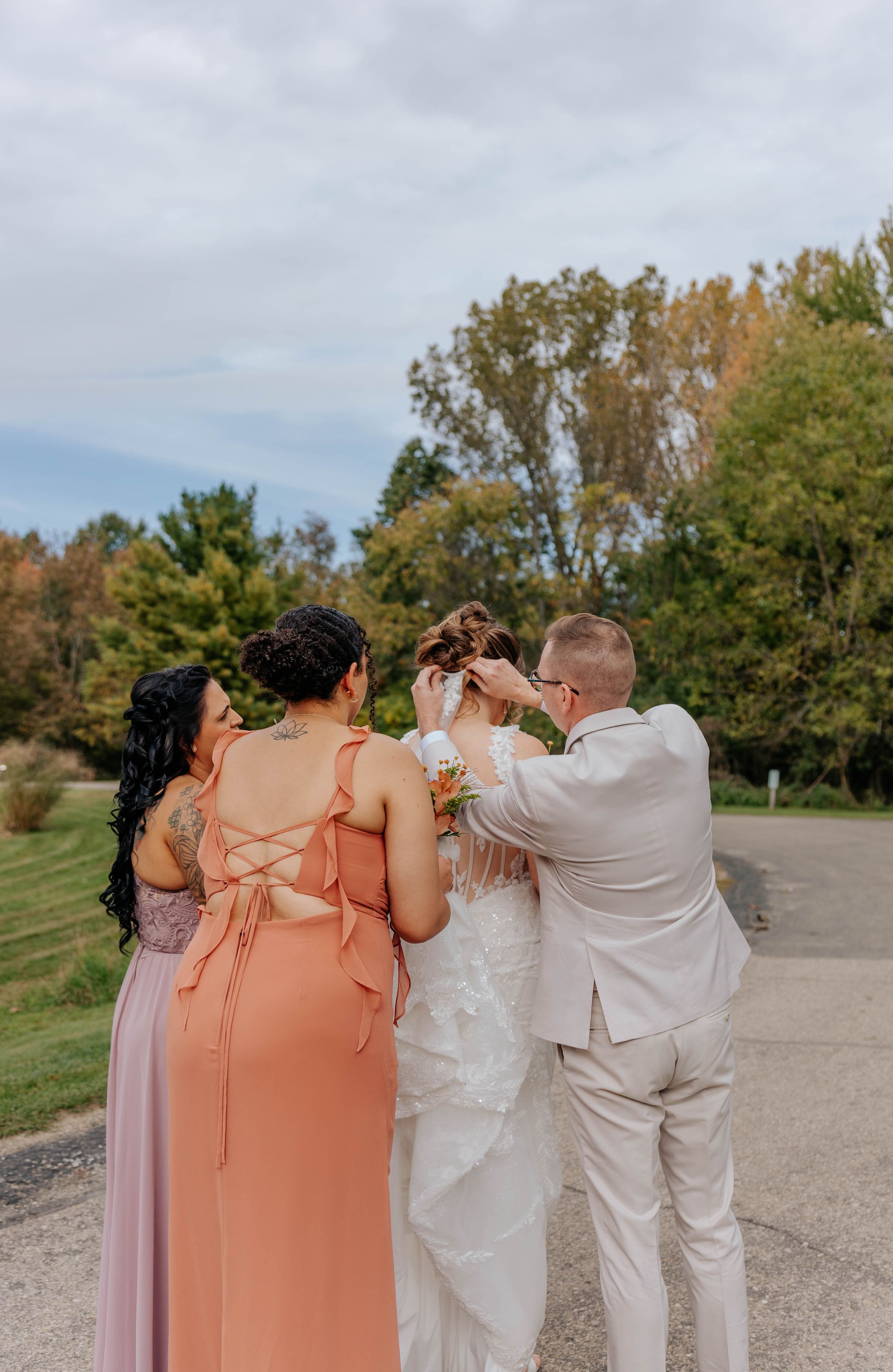 Bride surrounded by bridesmaids and groom adjusting her hair outdoors at a wedding ceremony.