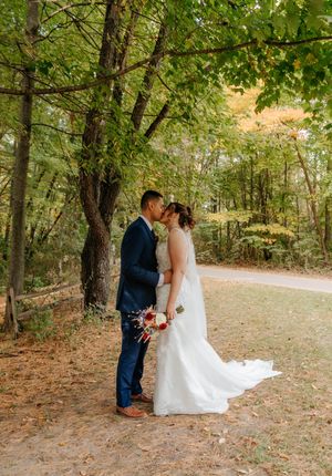 A couple shares a romantic kiss in a lush forest setting, the bride in a white gown holding a red bouquet.