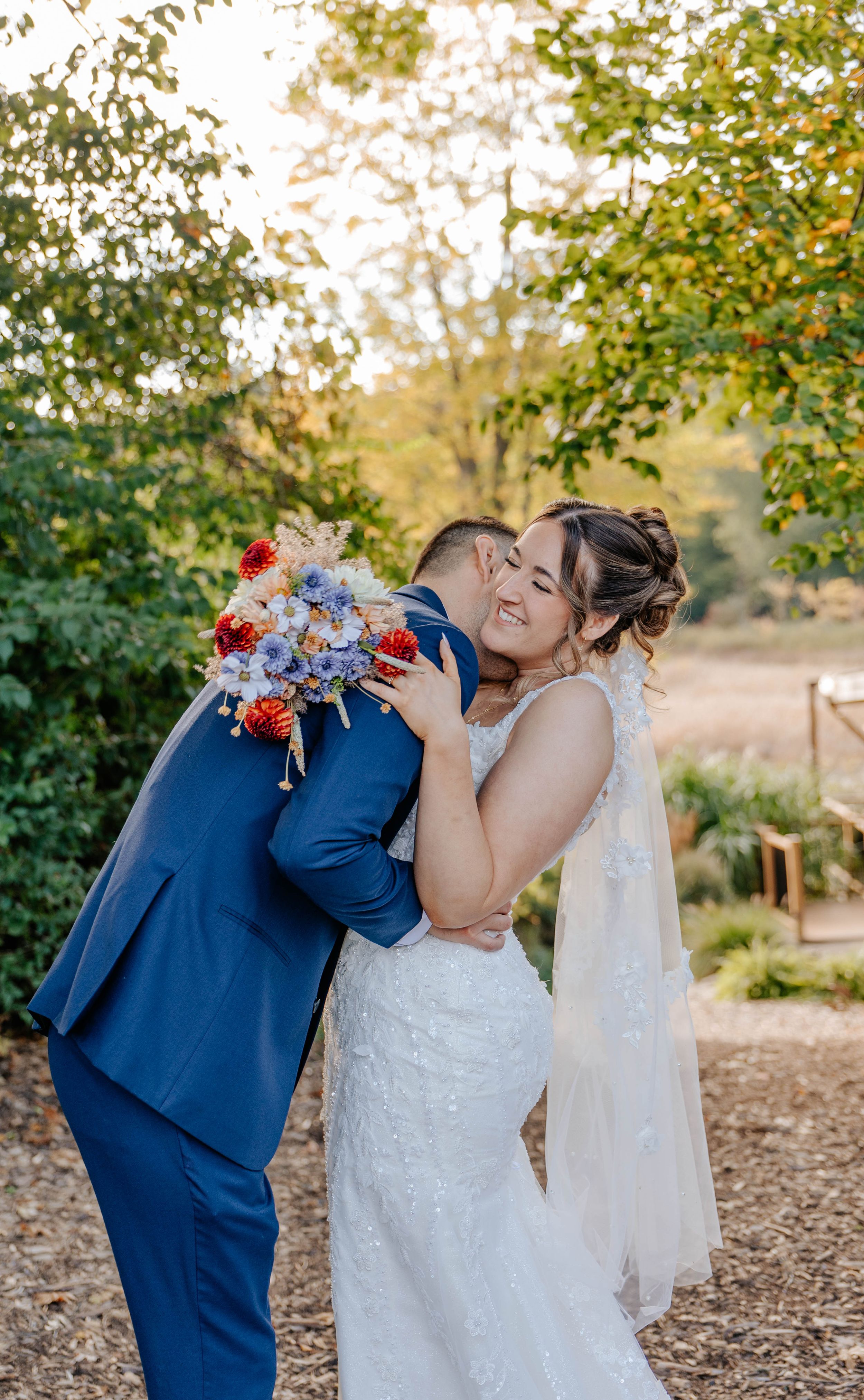 A bride in a white lace gown laughs as her groom in a blue suit kisses her cheek outdoors.