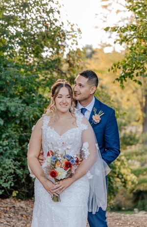Bride in lace gown holds colorful bouquet with groom in blue suit, posing outdoors among lush green trees.