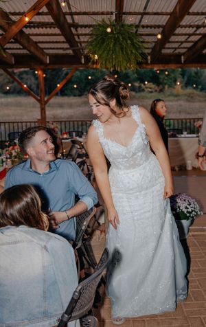 Bride in white lace gown smiling at seated guest during outdoor wedding reception with string lights.
