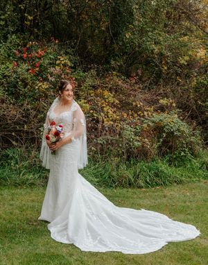 Bride in white lace mermaid gown with veil holds colorful bouquet, standing on green lawn with autumn foliage backdrop.