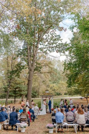 Outdoor wedding ceremony in a wooded setting with guests seated in white chairs facing the couple at a rustic wooden arch.