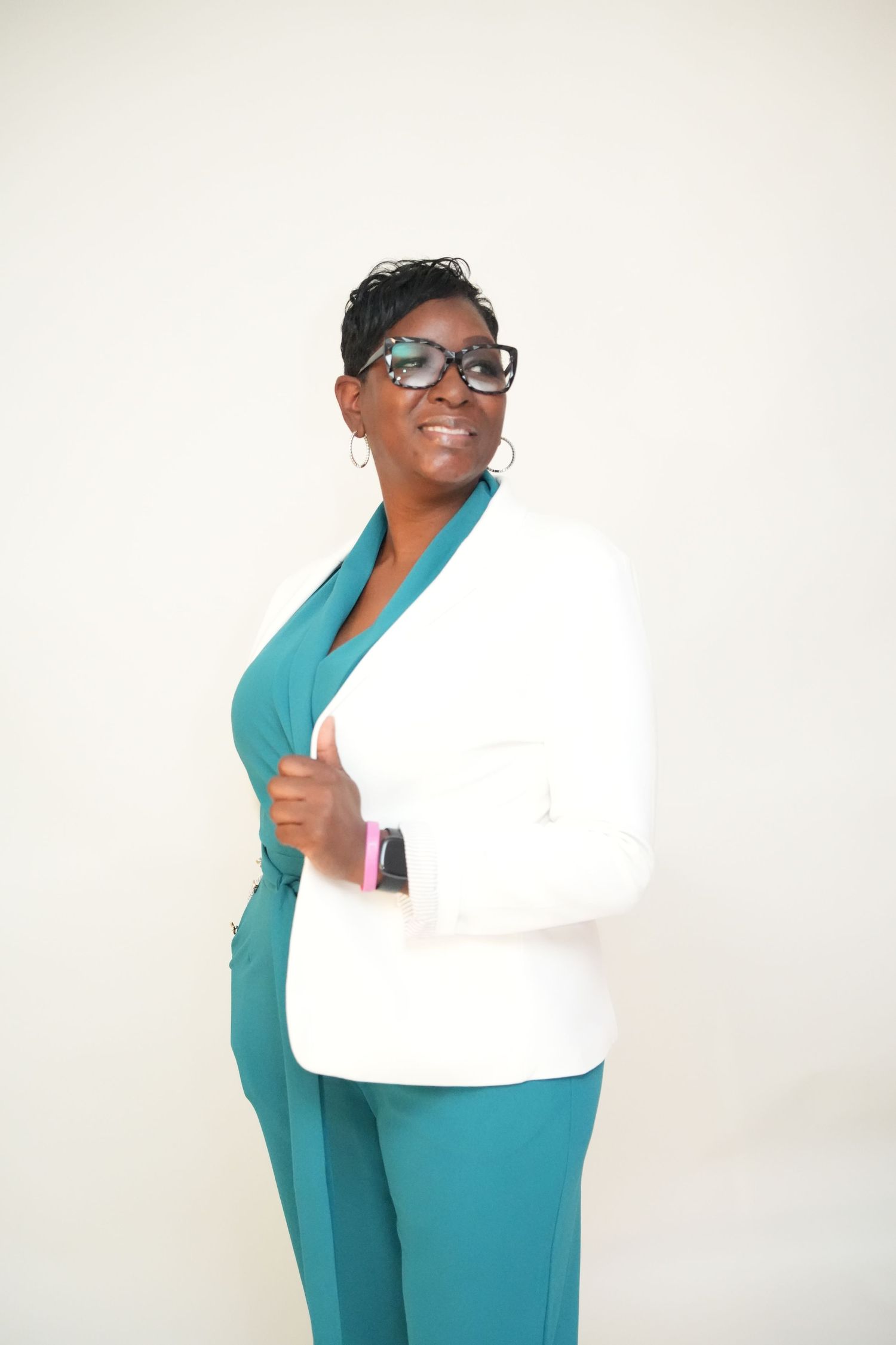 Smiling woman in glasses and hoop earrings poses in a white blazer over a teal jumpsuit against a plain light background.