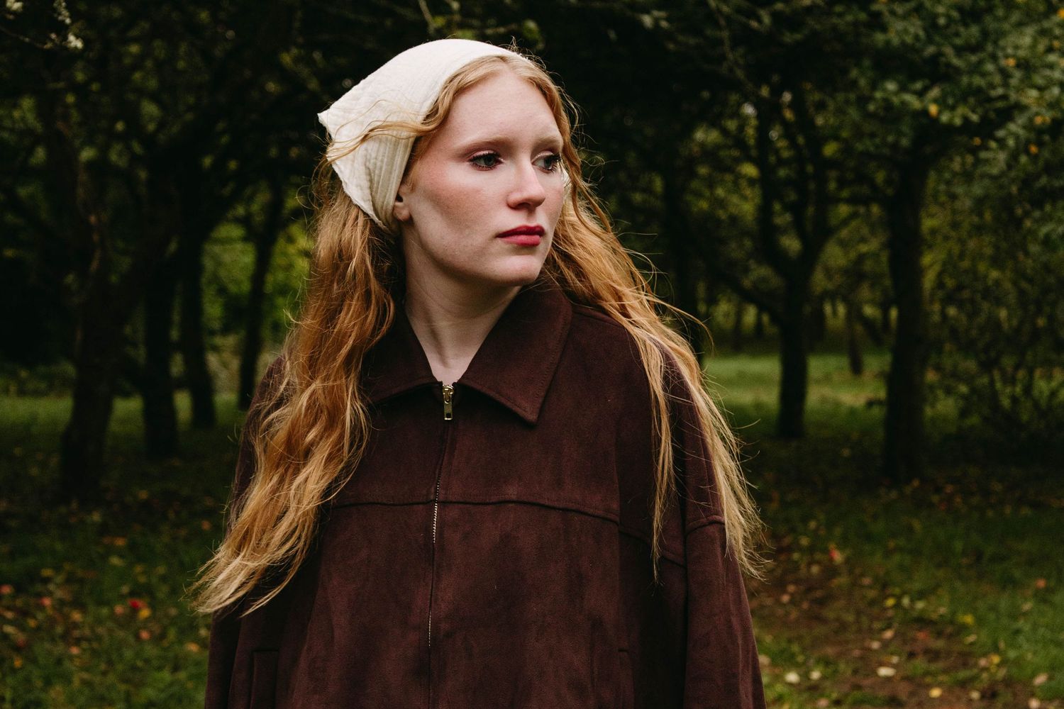 Girl with long auburn hair in a brown jacket and white lace skirt holds a red apple in a sunlit autumn park, wearing black knee-high socks.