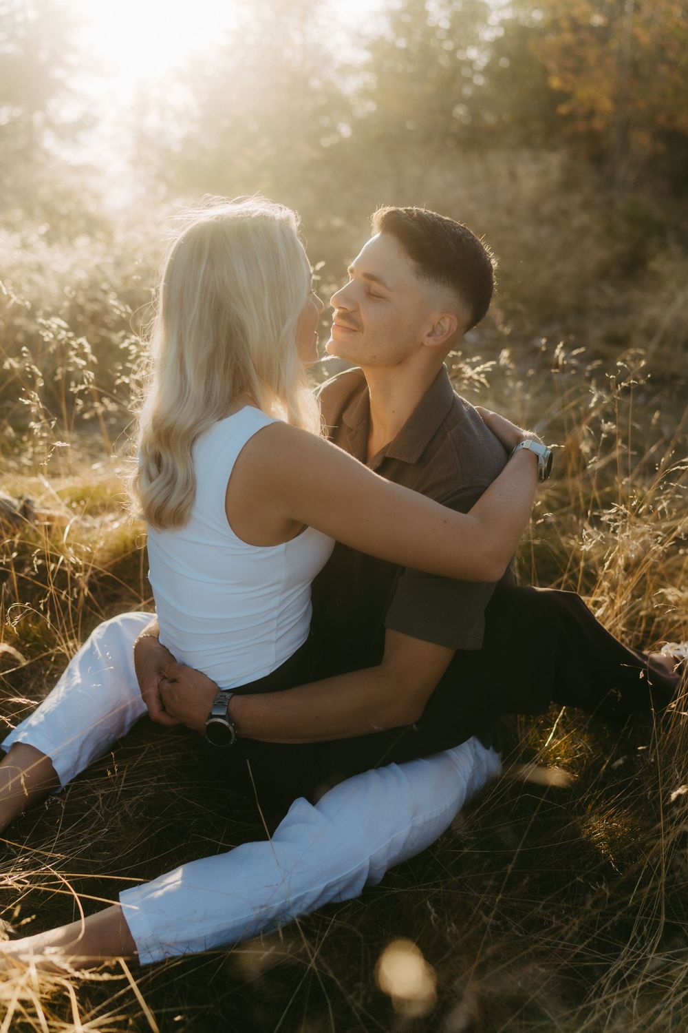 Couple sitting close together in tall golden grass, embracing and smiling during a warm sunset engagement photo session outdoors.