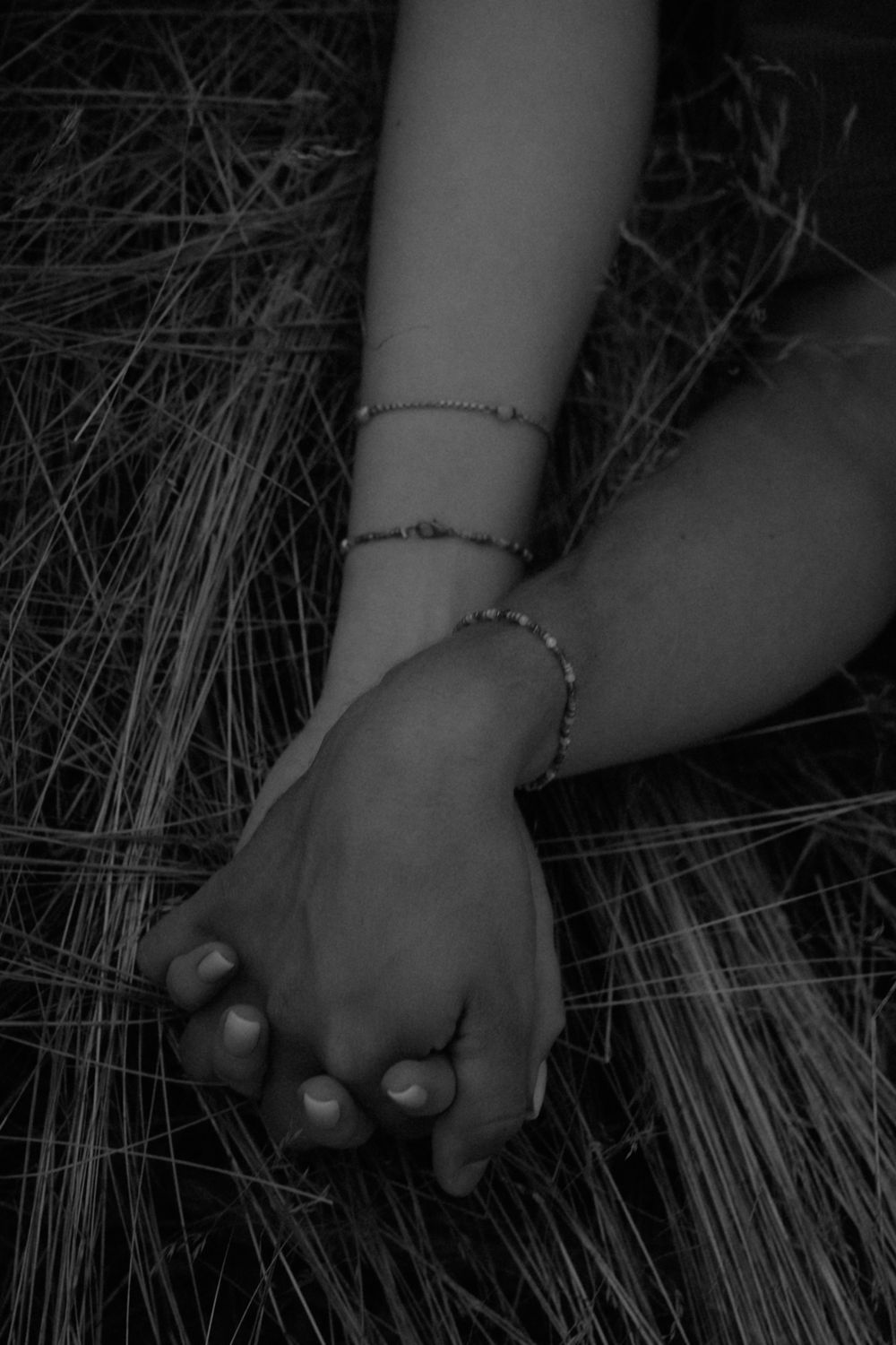 Black and white close-up of two hands clasped together on dry grass, both wrists wearing delicate beaded bracelets.