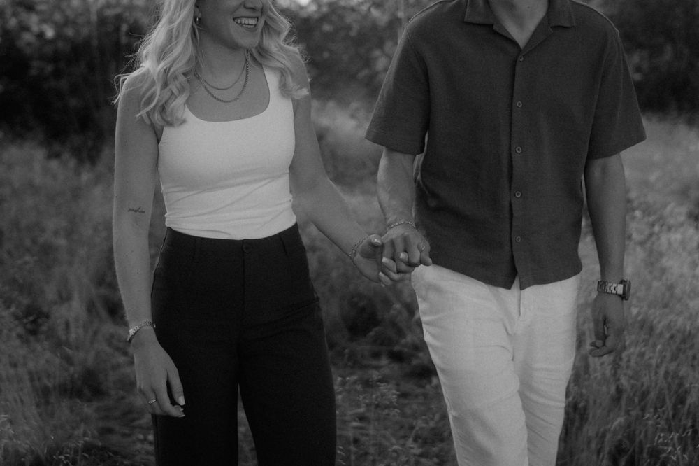 Black and white photo of a smiling couple holding hands while walking through a grassy field, she in a tank top and he in a button-up shirt.